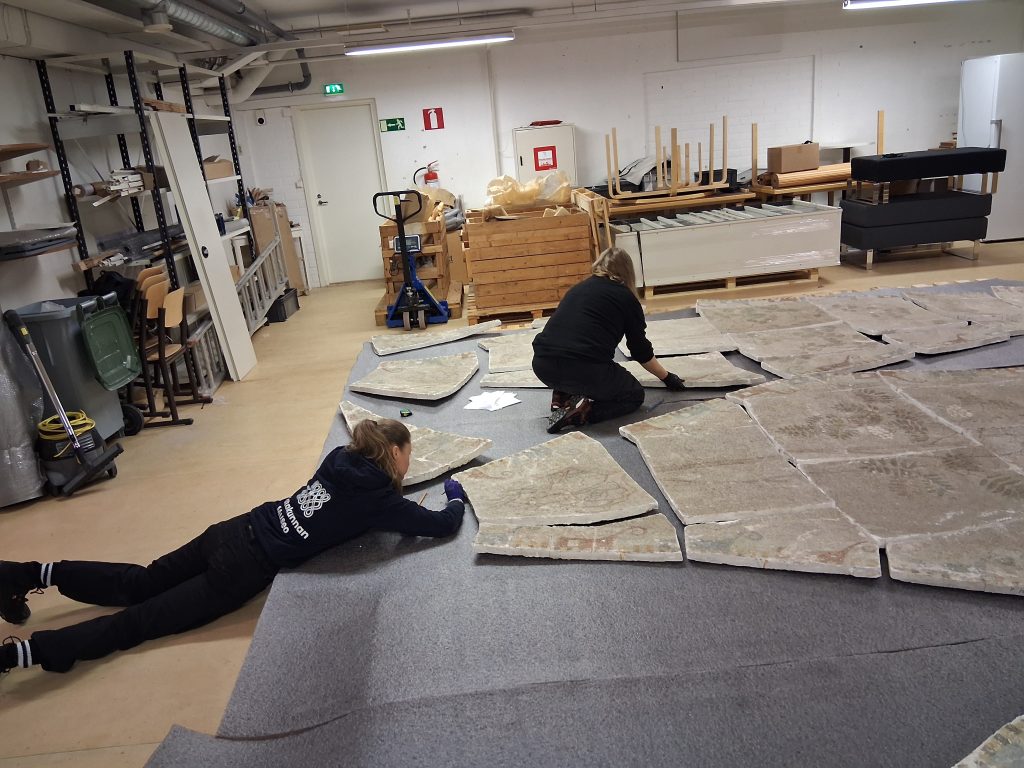 Kaksi henkilöä työskentelemässä museon varastotilassa freskon kappaleiden parissa.