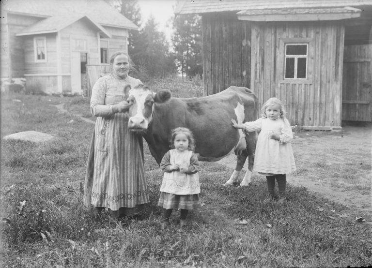 Nainen, kaksi lasta ja lehmä katsoo kameraan vanhojen puurakennusten edessä.
